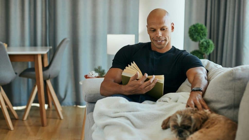 A man in a black t-shirt sits on a sofa, reading a book and petting a dog.