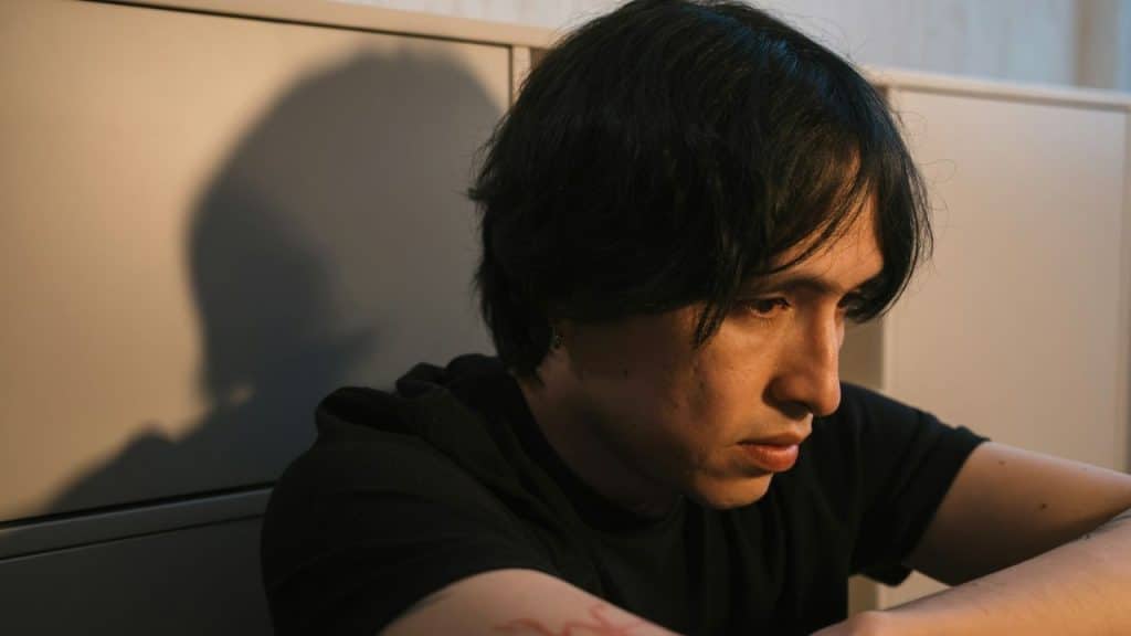 A man with dark hair in a black shirt looks downward while sitting in a dimly lit room.