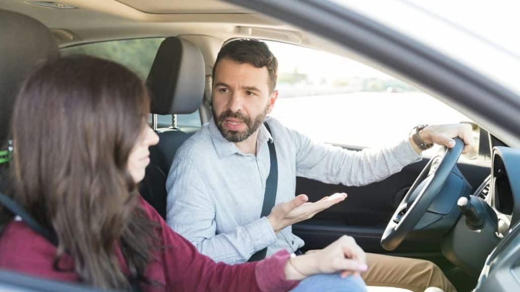 A bearded man drives a car and gestures while talking to a woman in the passenger seat.