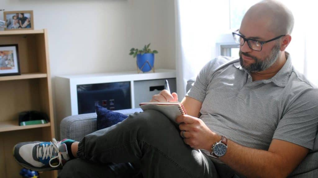 A man sits on a couch writing in a notebook.