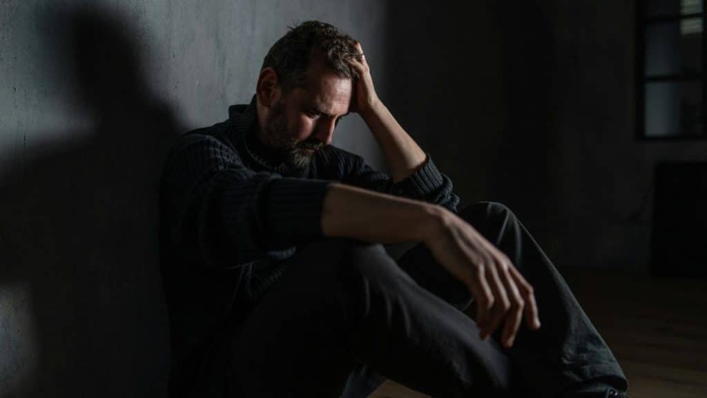 A man in a dark sweater sits on the floor against a wall, head down.