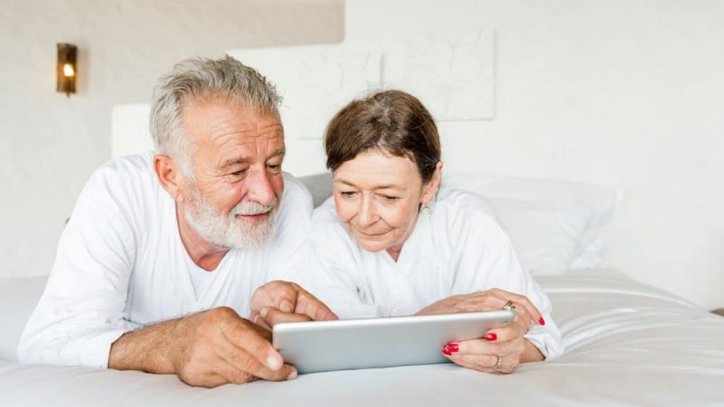 An elderly couple lies in bed, looking at and pointing at a tablet.