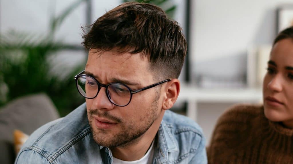 A concerned man with glasses looks away from a woman sitting nearby.