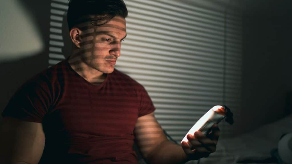 A man in a red shirt looking at a bright phone in a darkened room with blinds.