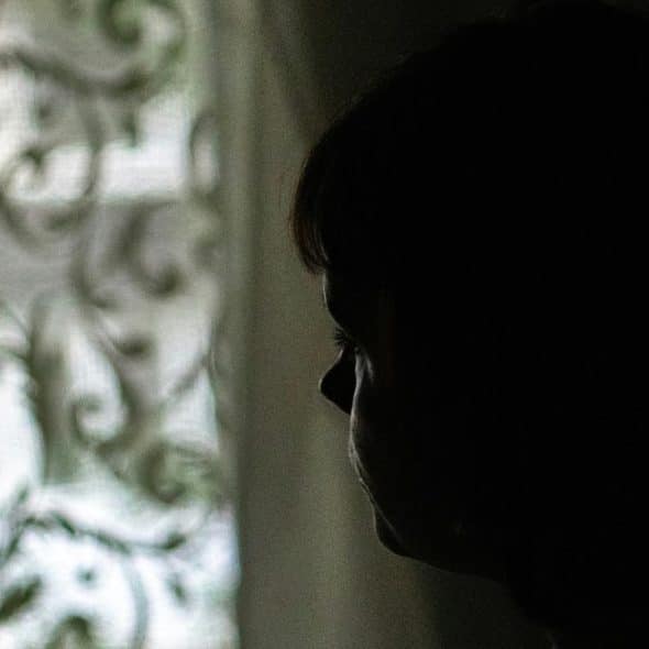 A silhouetted person gazing toward a softly lit window with a patterned curtain.