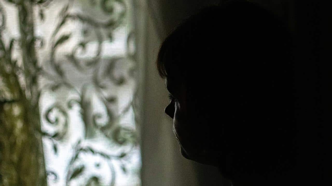 A silhouetted person gazing toward a softly lit window with a patterned curtain.