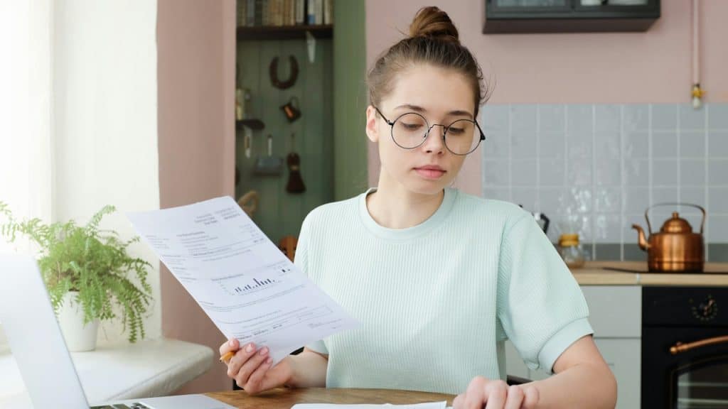 A young woman with glasses sits at a kitchen table, focused on reviewing financial documents