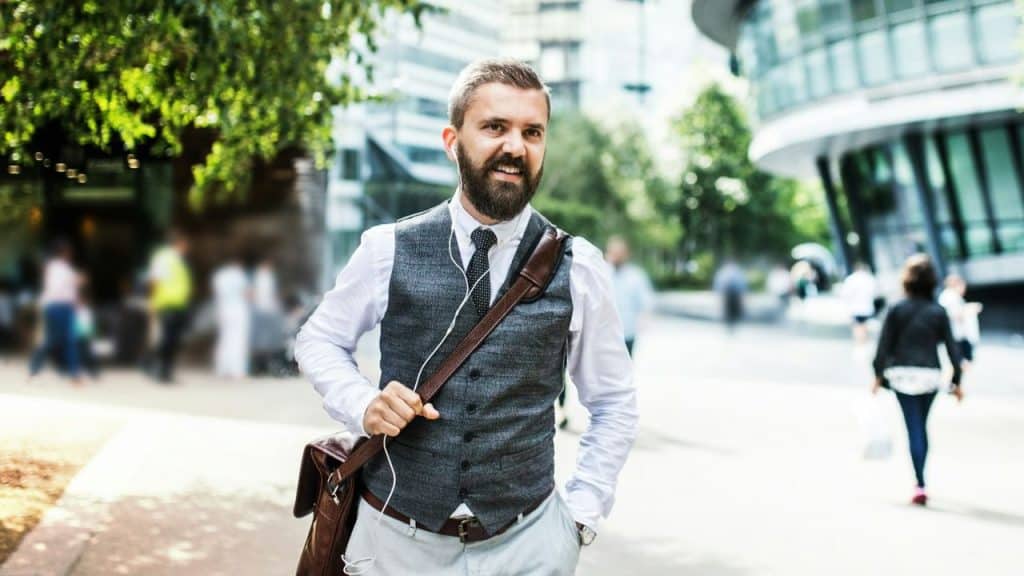 A bearded man in a grey vest and tie walks outdoors while smiling.