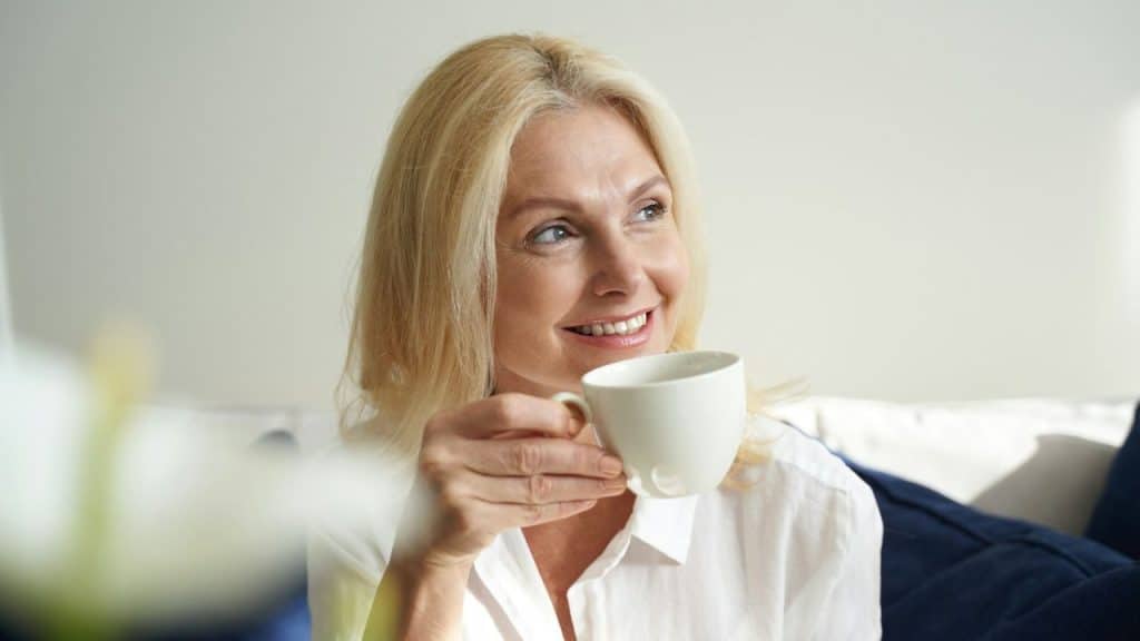 A smiling blonde woman holding a white mug and looking away against a neutral background.
