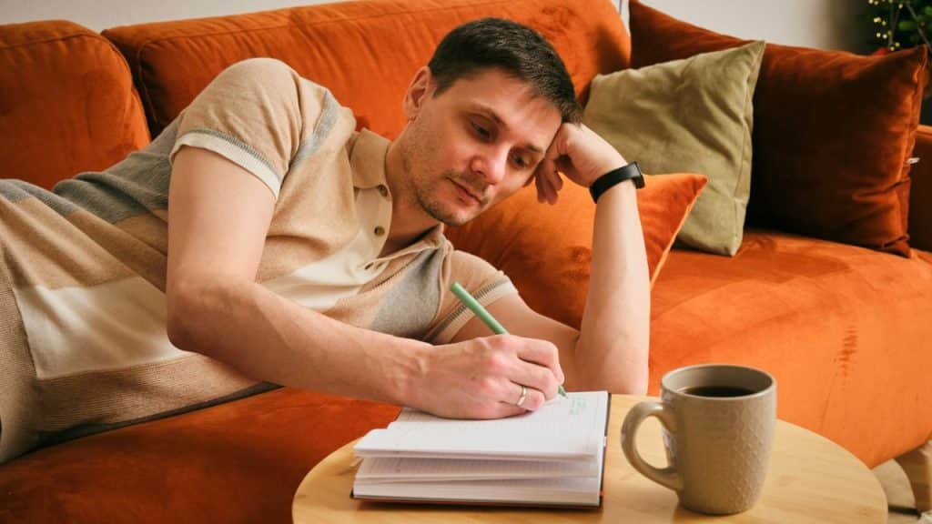 A man lying on an orange sofa while writing in a notebook next to a mug.