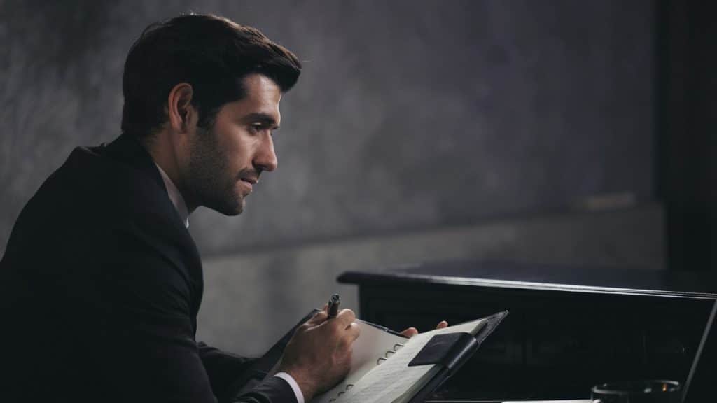 A man in a dark suit sits at a desk while writing in a leather-bound notebook.