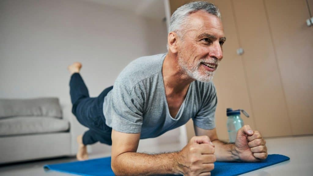 A gray-haired man smiling while performing a plank exercise on a blue yoga mat.