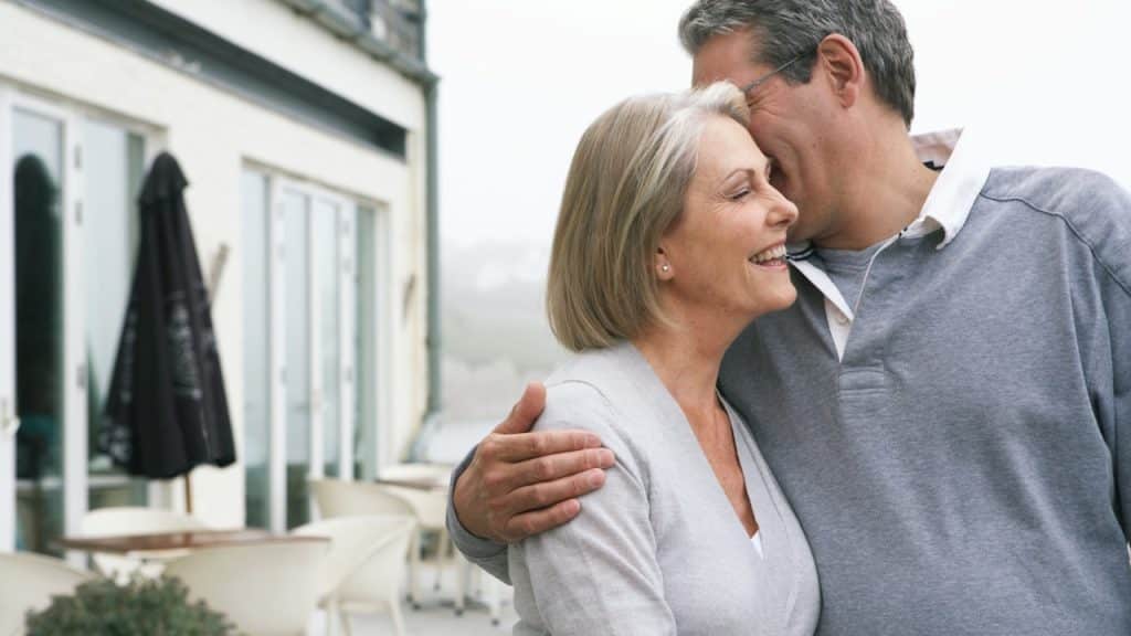 A smiling mature couple embracing warmly on an outdoor terrace, with the man leaning in toward the laughing woman.