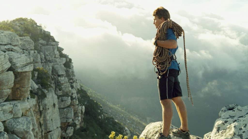A man with climbing rope over his shoulder stands on a rocky mountain peak.