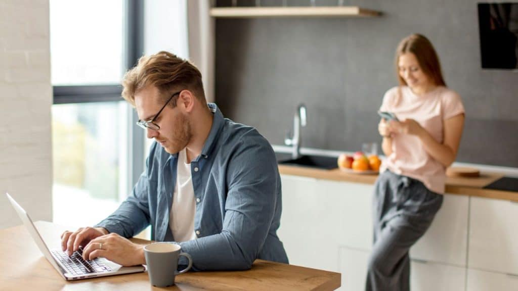 A man focused on his laptop while a woman looks at her phone in the background.