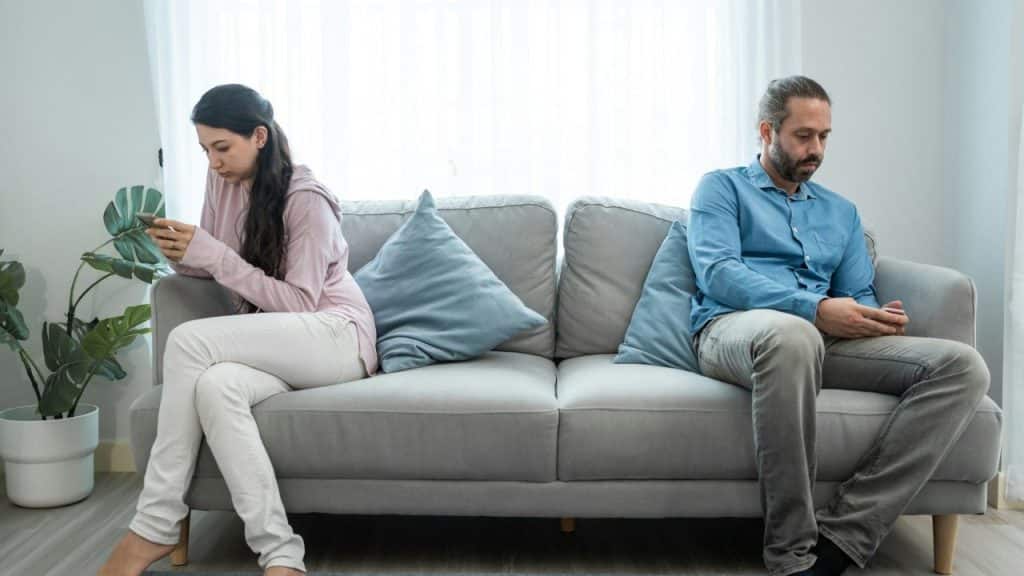A man and woman sit at opposite ends of a gray sofa, each using a smartphone.