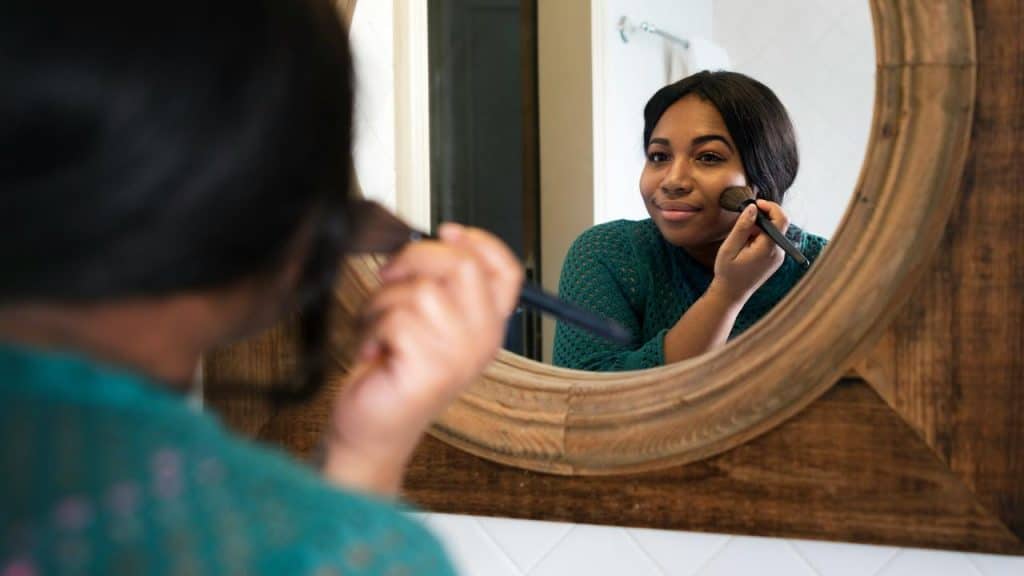 A woman applying makeup while looking in a bathroom mirror.