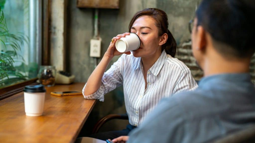 A woman in a striped shirt drinks from a white cup while sitting across from a man.