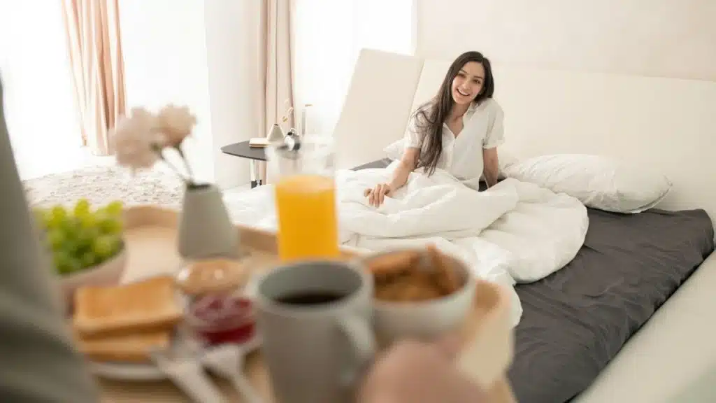 A person holds a tray of breakfast food toward a woman sitting up in bed.