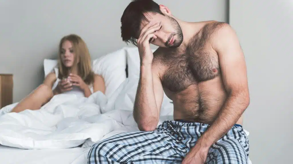 A shirtless man sits on a bed looking distressed while a woman watches him.