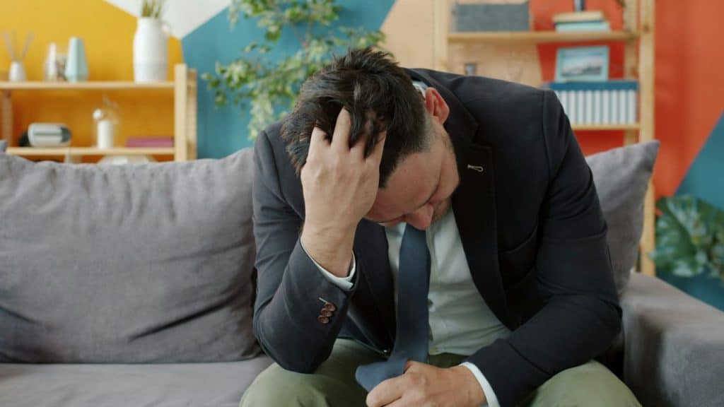 A man in a suit jacket sits on a couch with his hand on his head.