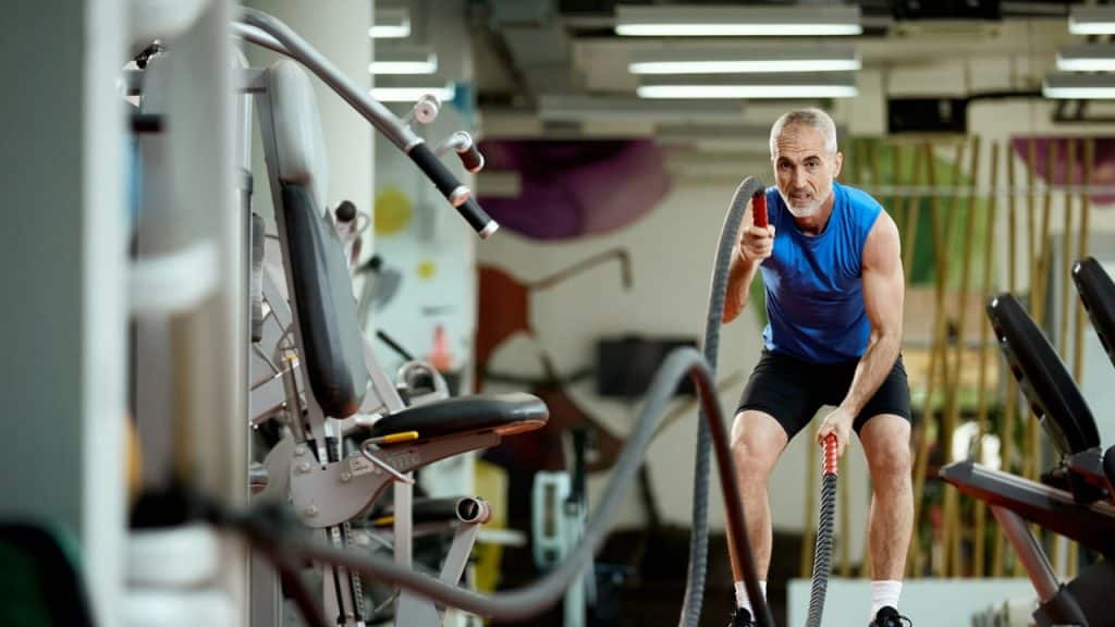 A man in a blue shirt uses heavy battle ropes while exercising inside a gymnasium.