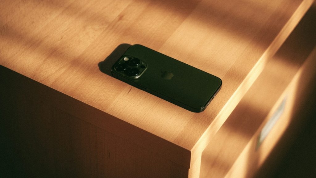 A black smartphone sits on a light-colored wooden surface in warm, natural light.