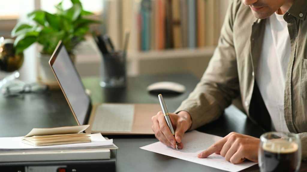 A person writes on a calendar with a pen at a desk with a laptop.