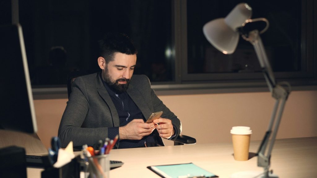 A man in a suit sits at a desk at night looking at his phone.