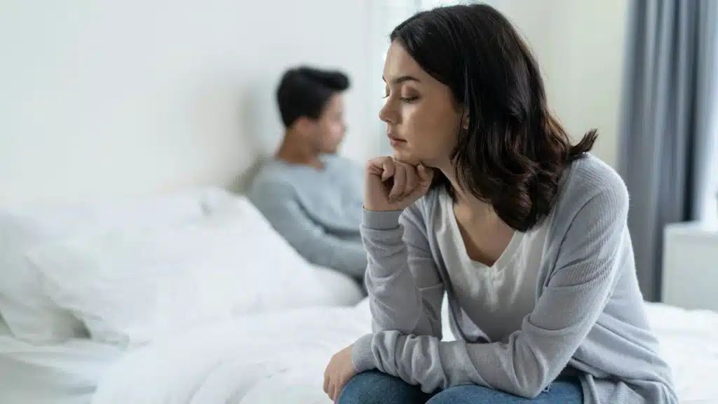 A woman is sitting on the bed while her angry husband sits on the other side of the bed.