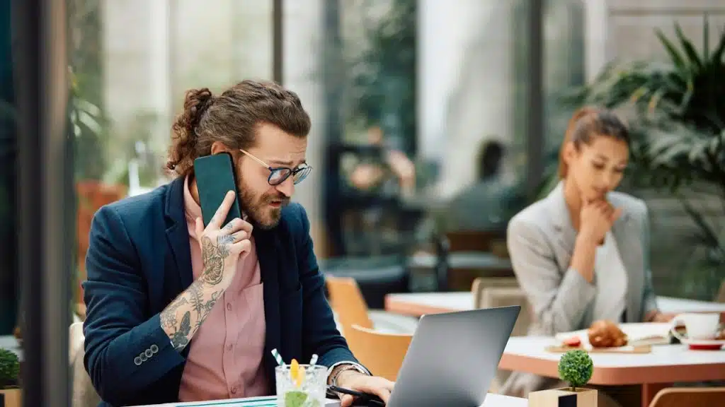 A man is talking on the phone while working on a laptop, while his female colleague is sitting behind him.