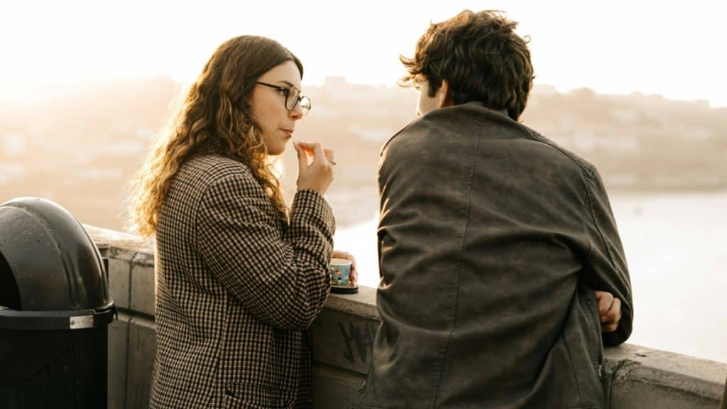 A man and woman lean against a concrete wall outdoors while talking at sunset.