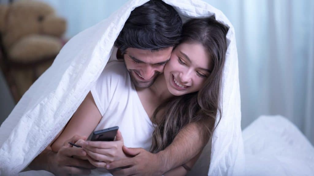 A happy couple cuddles under a white blanket, looking at a phone.