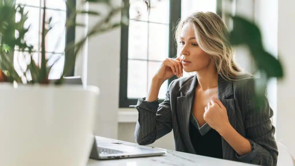 A woman in a blazer sitting at a desk and looking thoughtfully at her laptop.