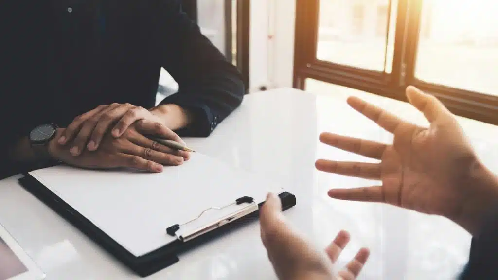 A person holding a pen over a blank clipboard while another gestures across a table.