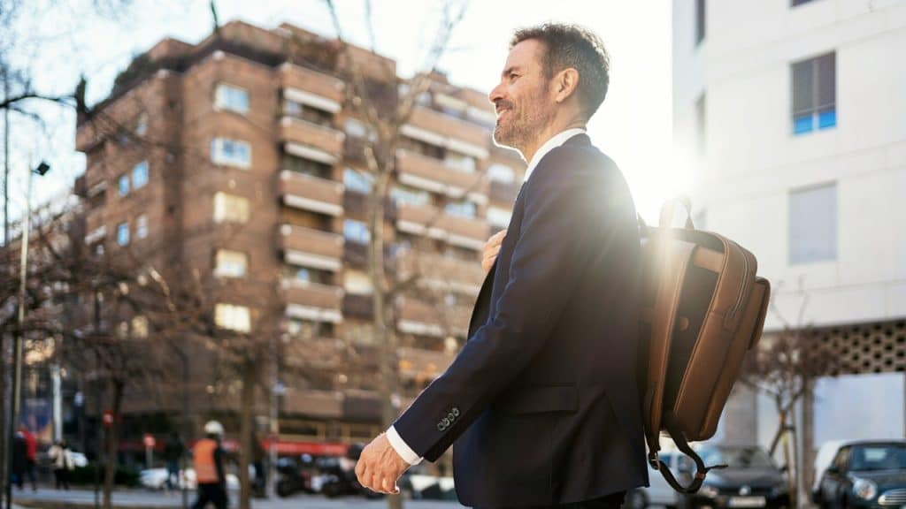 A smiling man in a suit walks through a city street carrying a brown leather backpack.