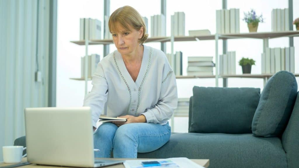 A woman in a white blouse and jeans sits on a sofa, looking at a laptop.