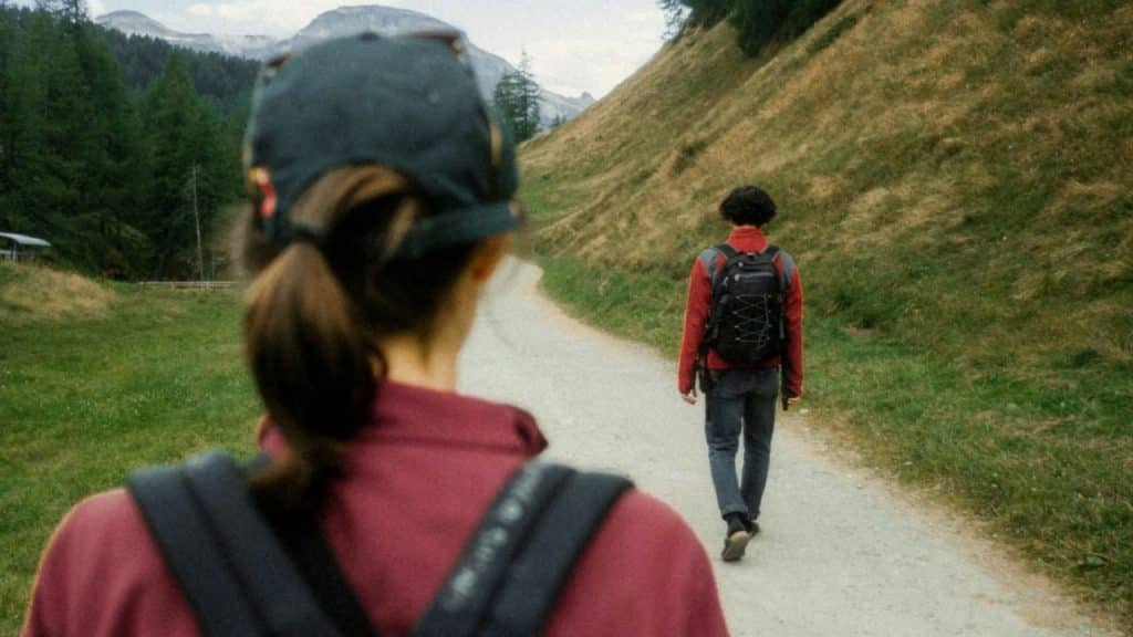 A woman with a backpack walks behind a man on a dirt path in the mountains