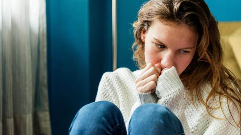 A young woman sitting curled up indoors looking anxious.