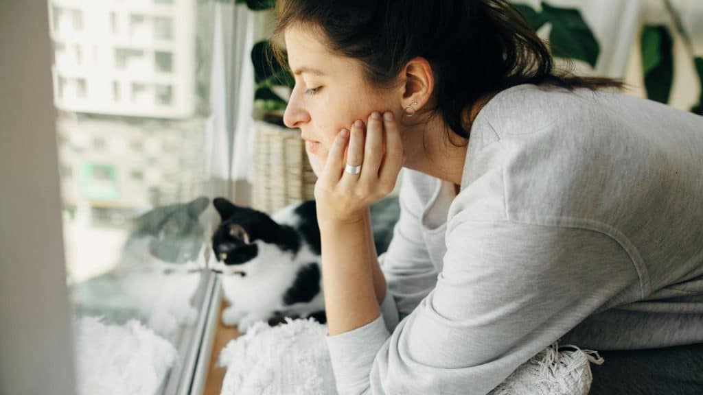 A person resting their chin on their hand while sitting by a window with a cat nearby.