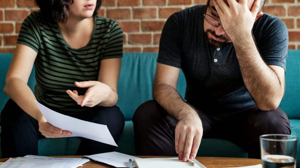 A woman gestures toward papers while a man sits beside her with his hand on his head.