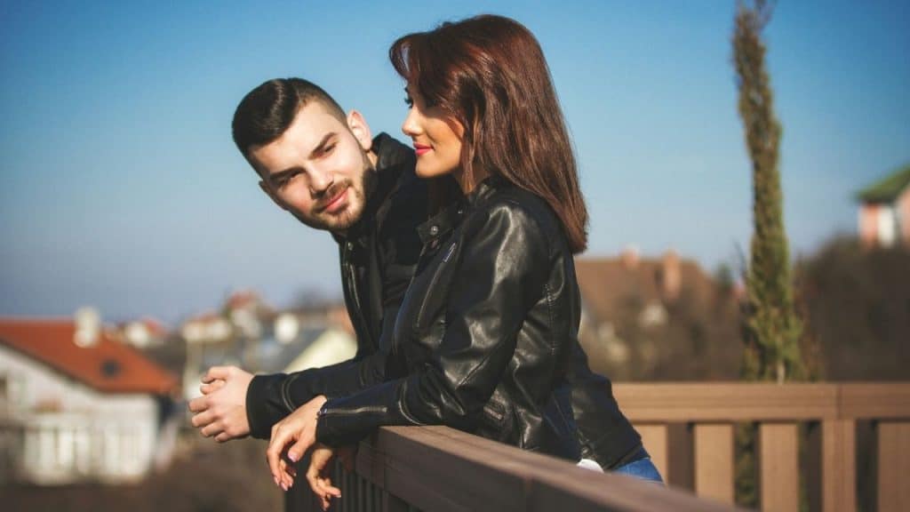 A man and woman in black leather jackets lean on a railing together outside.