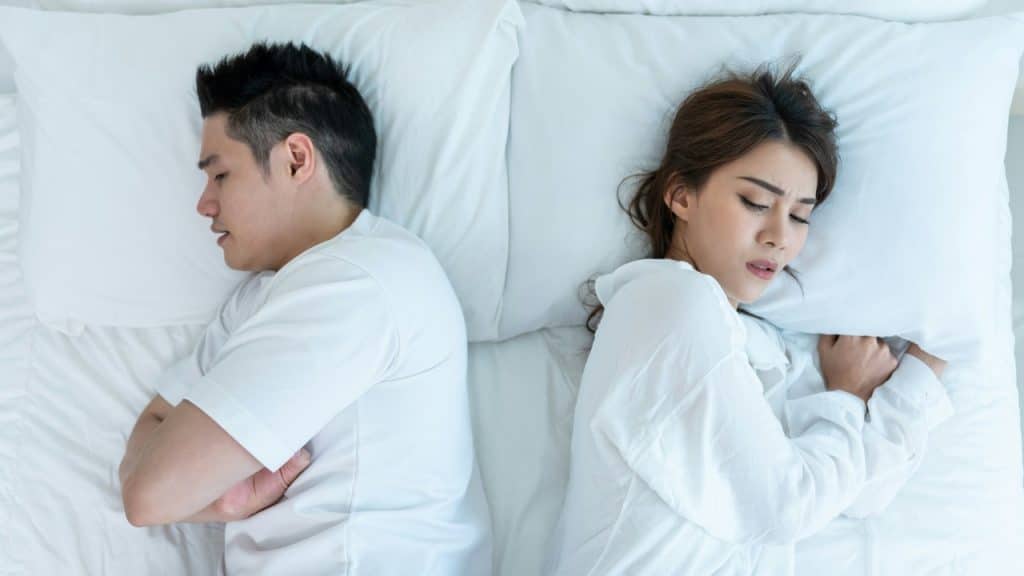 A man and woman lie in bed facing away from each other with frustrated expressions.
