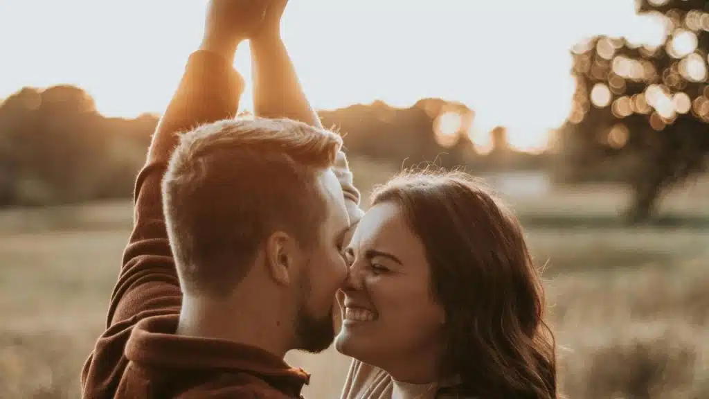 A joyful couple touching noses while raising their arms outdoors at sunset.