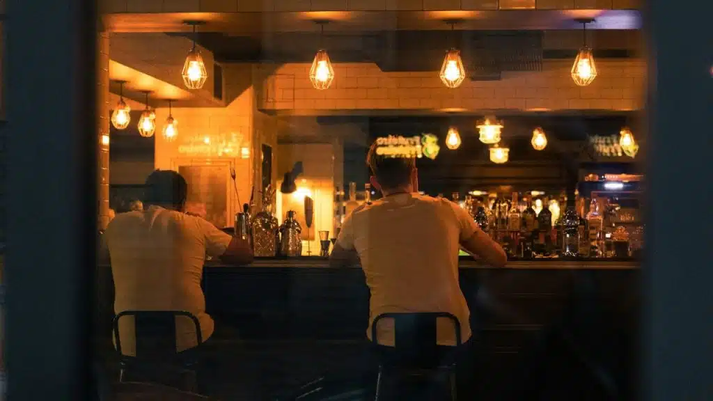 Two men in white shirts sit at a dimly lit bar, seen from outside.