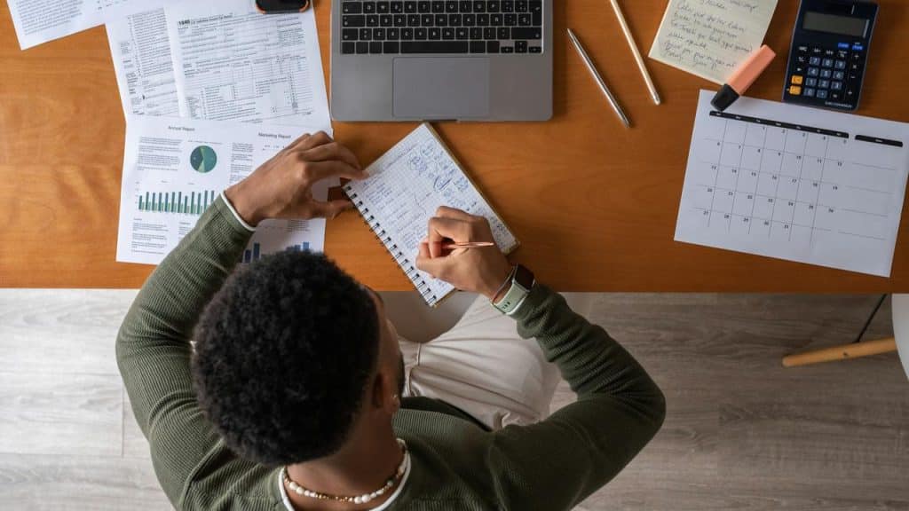 A person writing notes at a desk surrounded by documents, a laptop, and a calendar.