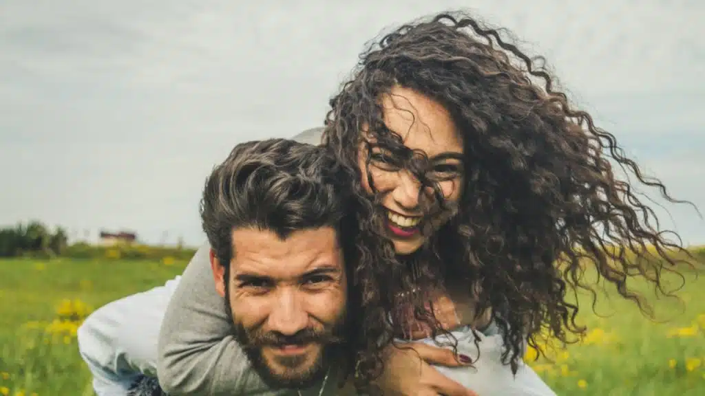 A bearded man carring a curly haired smiling woman on his back as they stand in a field outdoors.