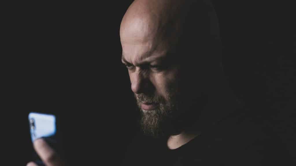 A man with a beard looks down at a glowing smartphone screen in a dark room.