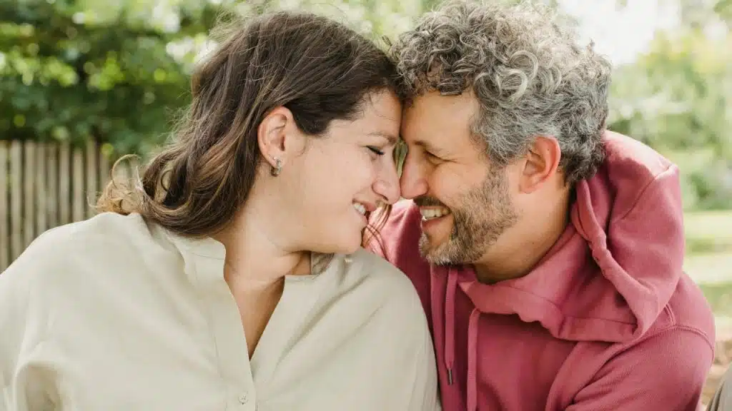 A man and woman smiling with their foreheads touching against a soft green outdoor background.