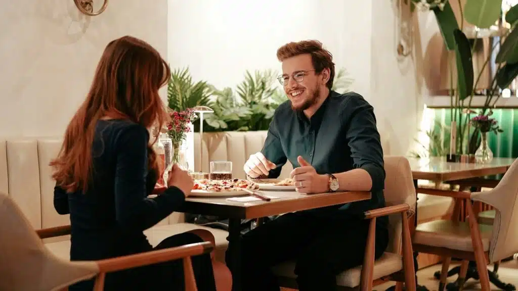 A smiling man with glasses sits at a restaurant table across from a woman with long hair.